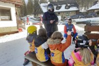 Policjant rozmawia z dziećmi-narciarzami podczas akcji profilaktycznej pod stacją narciarską. Na pierwszym planie widać dzieci, które zgłaszają się do odpowiedzi.