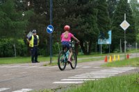 Dziewczyna jedzie rowerem po trasie egzaminu na kartę rowerową. W tle stoi policjant ruchu drogowego.
