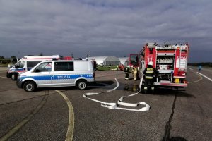 Strażacy podczas akcji ratowniczej, w trackie szkolenia na lotnisku, na drugim planie pojazdy służb, Straży Pożarnej i Policji.