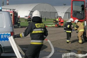 Strażacy podczas akcji ratowniczej, w trackie szkolenia na lotnisku, na drugim planie pojazdy służb, Straży Pożarnej i Policji.