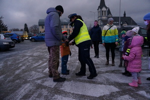 Policjantka zakłada dziecku opaskę odblaskową na ręke. Obok inne osoby dorosłe z dziećmi. Zdjęcie wykonane w dzień na parkingu.