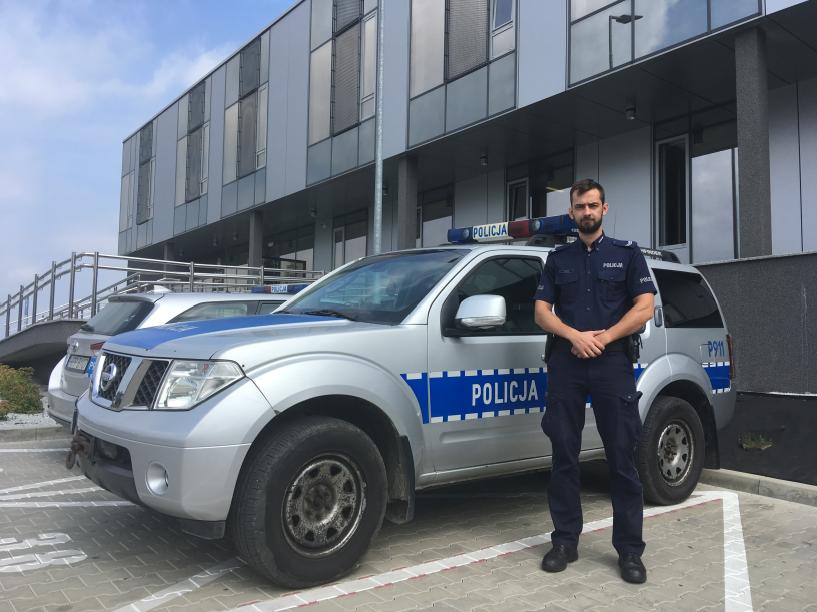 Policjant stoi przy radiowozie na tle budynku komendy