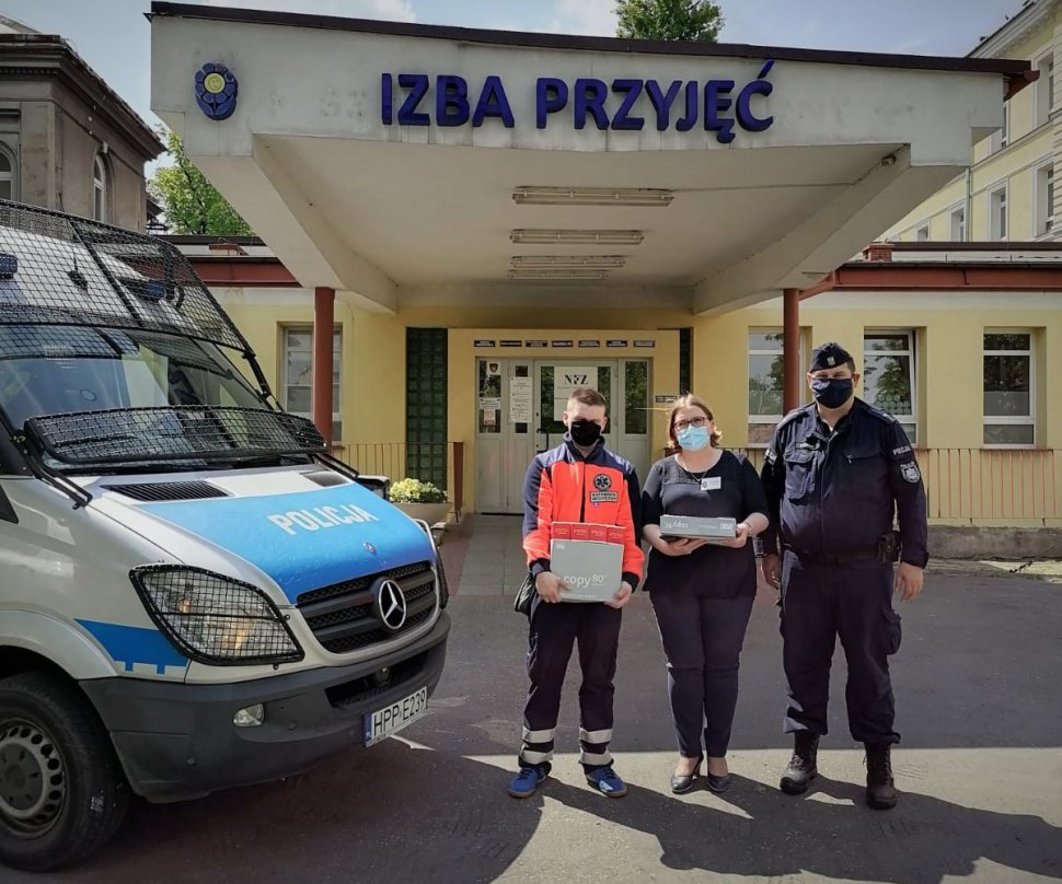 Policjant, ratownik medyczny i pielęgniarka stoją przed szpitalem. Obok widać maskę policyjnego radiowozu.