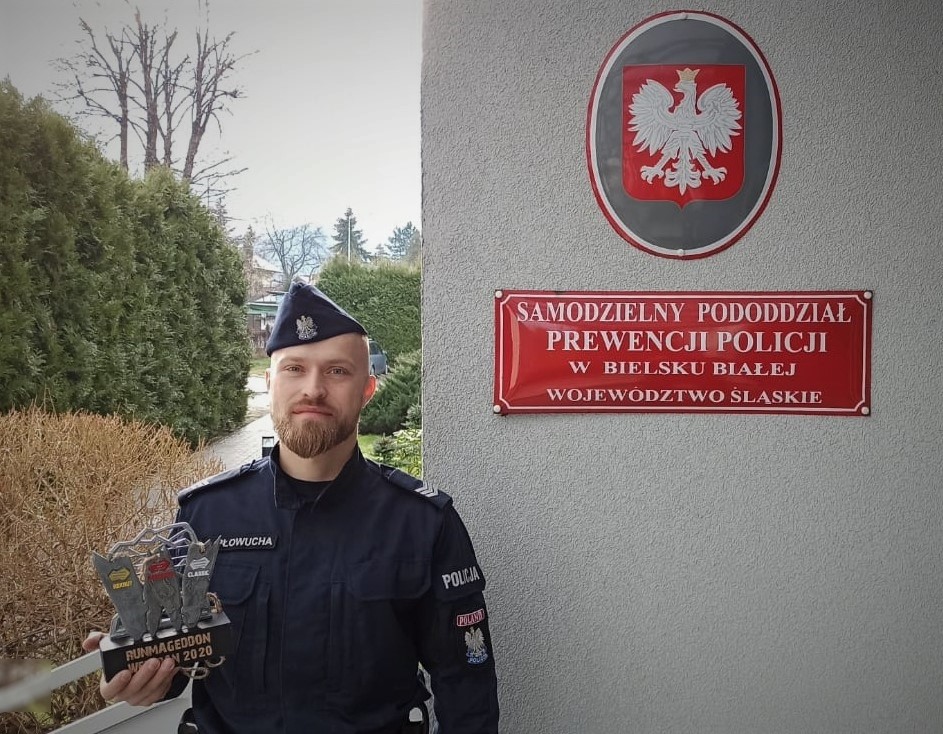 Sierżant sztabowy Jarosław Płowucha z Samodzielnego Pododdziału Prewencji Policji w Bielsku-Białej pozuje do zdjęcia w mundurze przed tablicą z nazwą jednostki i godłem Polski.