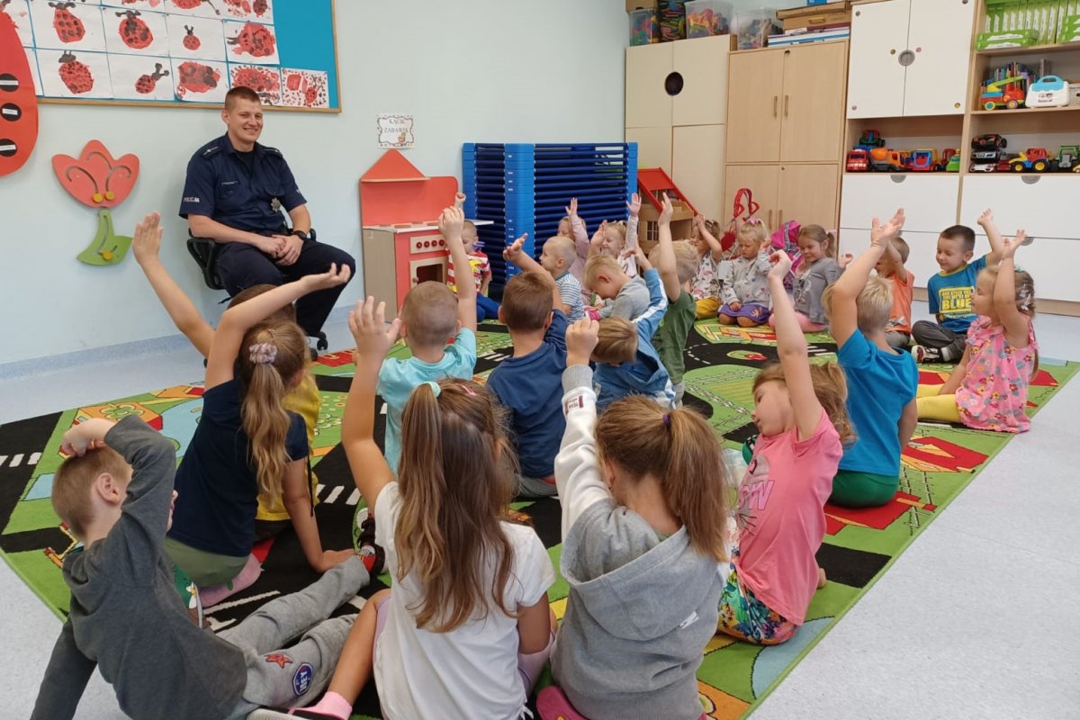 Dzieci zgłaszają się podnosząc rękę. Na drugim planie siedzi umundurowany policjant.