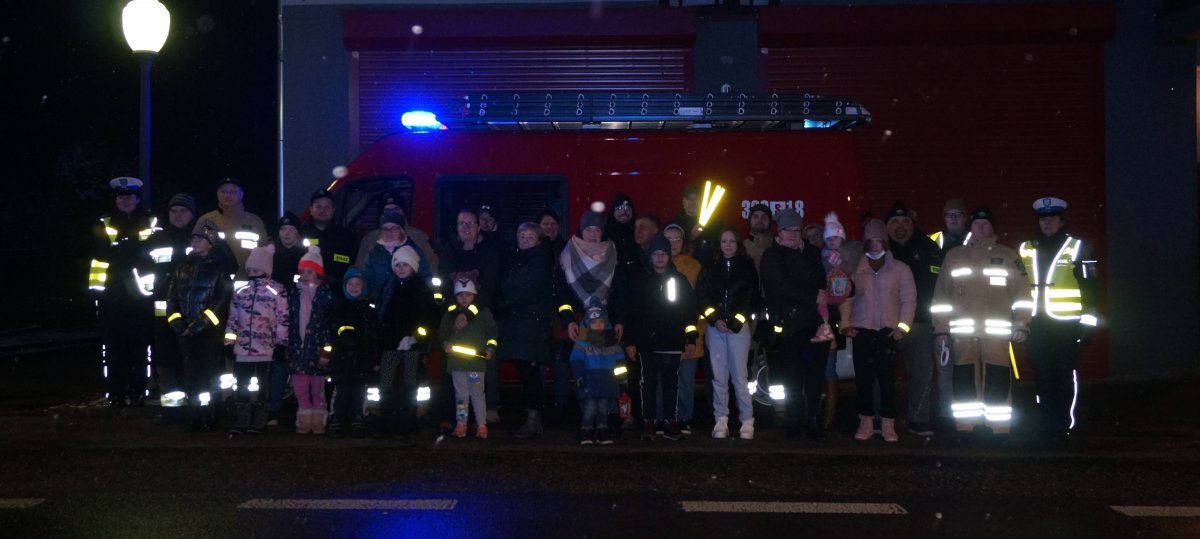 Uczestnicy akcji profilaktycznej, policjanci, strażacy, redaktorzy mediów na wspólnym zdjęciu pod siedzibą OSP. Zdjęcie wykonane na ulicy podczas akcji profilaktycznej po zmierzchu.