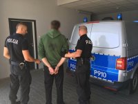 Policjant trzyma zatrzymanego. Drugi policjant otwiera drzwi radiowozu oznakowanego.