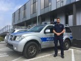 Policjant stoi na tle radiowozu przed budynkiem komendy.