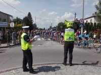 Policjant oraz marshall wyścigu stoją na ulicy, którą przejeżdża peleton kolarski.