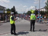 Policjant oraz marshall wyścigu stoją na ulicy, którą przejeżdża peleton kolarski.