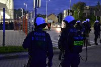 Policjant z miotaczem gazu stoi z drugim policjantem. W tle stadion piłkarski.
