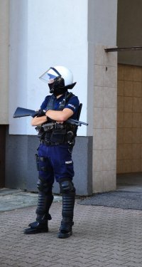 Policjant z bronią długą gładkolufową stoi przy wjeździe do kamienicy.