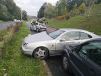 Trzy rozbite pojazdy stoją na jezdni, w tle służby ratownicze.