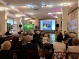 Seniorzy siedzą w sali konferencyjnej. W tle stolik prezydialny z przedstawicielami ZUS Bielsko-Biała i policjantem w mundurze i okno prezentacji z napisem; Nie daj się oszukać.