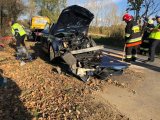 Wrak rozbitego pojazd stoi na jezdni, obok strażacy, policjant i pracownik pomocy drogowej.