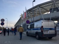 Policyjny radiowóz stoi zaparkowany w rejonie stadionu piłkarskiego. Chodnikiem przechodzą kibice.