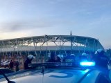 Policyjny radiowóz z włączonymi sygnałami błyskowymi. W tle policjanci oddziałów prewencji i stadion piłkarski.