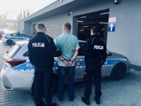 Policjanci trzymają zakutego w kajdanki mężczyznę. Stoją przed radiowozem.