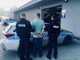 Policjanci trzymają zakutego w kajdanki mężczyznę. Stoją przed radiowozem.