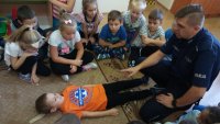 Policjant tłumaczy dzieciom udzielanie podstawowej pierwszej pomocy. Jedno z dzieci leży, pozostałe są zgromadzone wokół niego i prowadzącego zajęcia policjanta.