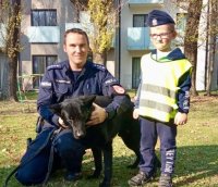 Policjant z psem, obok dziecko w kamizelce odblaskowej i policyjnej czapce.