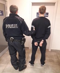 Policjant trzyma mężczyznę, z kajdanki założonymi na ręce z tyłu. Stoją w przedsionku korytarza komendy policji.
