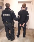 Policjant trzyma mężczyznę, z kajdanki założonymi na ręce z tyłu. Stoją w przedsionku korytarza komendy policji.