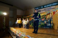 Policjantka stoi na scenie, trzymając odblaski, na drugim planie stoją dzieci.