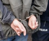 Mężczyzna prowadzony przez policjanta, na ręce trzymane z tyłu ma założone kajdanki.
