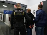 Policjant w mundurze i drugi w cywilu trzymają zatrzymaną, która ma założone kajdanki na ręce trzymane z tyłu.