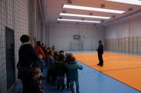 Policjant stoi z dziećmi na sali gimnastycznej. Rozmawiają o sporcie.