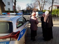 Policjant i ksiądz rozmawiają z przechodniami. Stoją przy samochodzie policyjnym.