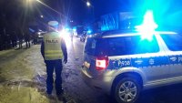 Policjant z ruchu drogowego zabezpiecza przejazd. Stoi obok radiowozu.