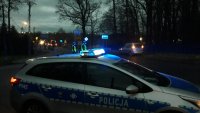 Radiowóz policyjny z włączonymi sygnałami świetlnymi. Na drugim planie policjanci kierują ruchem.