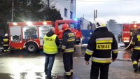 Policjant i strażacy, obok wozy strażackie. Na drugim planie budynek, z którego wydobywa się dym.
