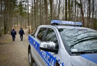 Policjanci idą leśną drogą, widok przez maskę policyjnego wozu.