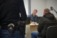 Na pierwszym planie widać policyjny pistolet przy pasku policjantki. W tle policjant kryminalny rozmawia z zatrzymanym.