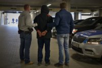 Policjanci ubrani po cywilnemu stoją z zatrzymanym, który ma założone kajdanki na ręce trzymane z tyłu. Obok radiowóz policyjny.