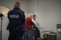 Technik kryminalistyki pobiera od zatrzymanego odciski palców. Na pierwszym planie policjant w mundurze dozoruje zatrzymanego.