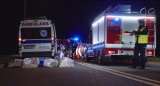 Policjant wskazuje latarką kierunek ruchu. W tel wóz strażacki i karetka pogotowia.