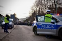 Policjantka stoi przy otwartych drzwiach radiowozu. Obserwuje tłum idący ulicą.