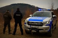 Policjant i Strażnik Leśny na leśnej drodze stoją obróceni tyłem przy radiowozie.