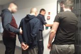 Policjanci doprowadzają zatrzymanego. Stoją przed kratą policyjnego aresztu.