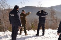 Strażnik leśny wskazuje miejsce na zboczu wzniesienia. Policjant spogląda przez lornetkę.