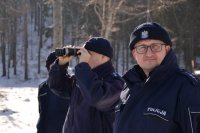Policjanci stoją, jeden z nich spogląda przez lortnetkę.
