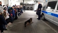Dzieci stoją w szeregu na podziemnym parkingu komendy. Policjant prezentuje umiejętności policyjnego psa.