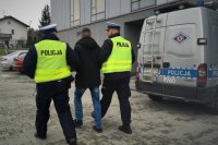 Policjanci z ruchu drogowego prowadzą zatrzymanego, obok stoi radiowóz policyjny.