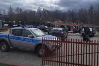 Policyjny oznakowany radiowóz stoi na terenie skupy auto-części, w tle nieumundurowani policjanci prowadzą czynności.