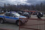 Policyjny oznakowany radiowóz stoi na terenie skupy auto-części, w tle nieumundurowani policjanci prowadzą czynności.