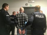 Umundurowani policjanci trzymają zatrzymanego zakutego w kajdanki.